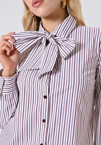 Camicia bianca con righe verticali bordeaux, dotata di un grande colletto a farfalla, chiusura con bottoni e maniche lunghe con dettagli sui polsini.