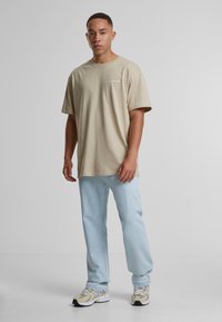 Beige, oversized póló rövid ujjal, kis logóval. Világoskék, egyenes szabású farmernadrág és bézs sneakers fehér részletekkel.