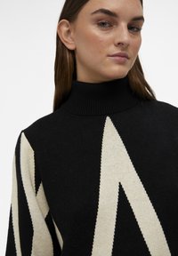 Pull noir et crème avec un col roulé haut, un motif géométrique en zigzag et un tissu tricoté texturé. La vue rapprochée met en valeur les détails du design.