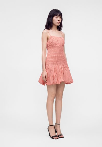 Femme debout portant une mini-robe rose froncée sans manches avec de fines bretelles et des sandales noires à talons hauts sur un fond uni.