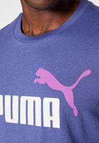 Violets kokvilnas T-krekls ar lielu baltu "PUMA" logo un rozā lecošā pundura grafiku, apaļu kakla izgriezumu un īsām piedurknēm.