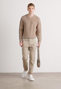 Hellbrauner Strickpullover mit V-Ausschnitt, beige schlanke Hose und weiße Sneakers. Hält eine graue Umhängetasche und steht vor einem neutralen Hintergrund.