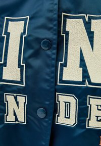 Modrá saténová bunda se velkými vyšívanými bílými písmeny 'I N D E', s lesklým modrým knoflíkem. Hladká textura s lesklým povrchem.