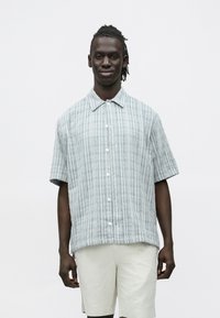 Mees heegeldatud juustega, seljas lühikeste varrukatega helesinine ruuduline nööbitav särk ja beežide lühikeste pükstega, seisab valge tausta ees.