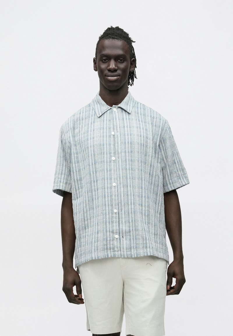 Mees heegeldatud juustega, seljas lühikeste varrukatega helesinine ruuduline nööbitav särk ja beežide lühikeste pükstega, seisab valge tausta ees.