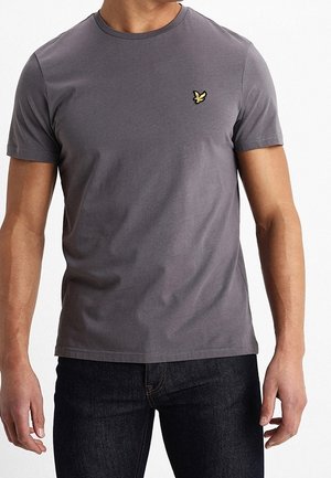 Mann trägt ein eng anliegendes dunkelgraues kurzärmeliges T-Shirt mit einem kleinen gelben und schwarzen Vogel-Logo auf der Brust und dunkle Jeans.