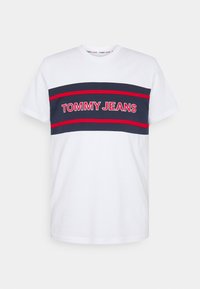 Vit bomulls-T-shirt med marinblå horisontella ränder. Har texten "TOMMY JEANS" i röd skrift över bröstet. Kort ärm, rundad halsringning.