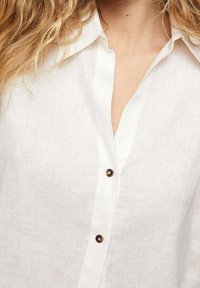 Kvinde iført en hvid linnedskjorte med skildpadde-knapper, krøllet blond hår synligt på venstre side.