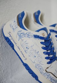 Zapatillas blancas con acentos azules presentan un diseño a lápiz de paisajes. El texto en el costado dice: “Tropezienne, mon amour.” Suela de goma texturizada.