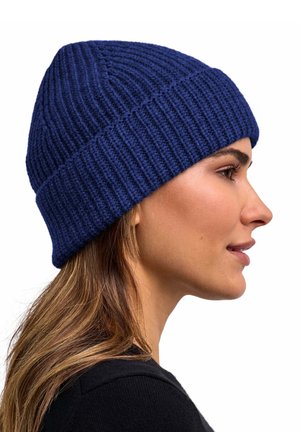 Vrouw met lang haar die een geribbelde, donkerblauwe gebreide muts draagt, kijkend naar rechts tegen een witte achtergrond.