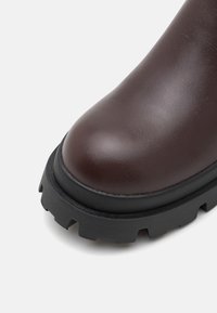 Bout noir en cuir brun foncé avec semelle épaisse à crampons noirs, conçu pour la durabilité et la traction.