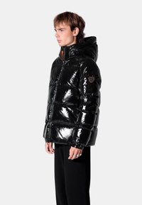 Doudoune noire brillante avec capuche, dotée d'une fermeture éclair à l'avant, de poignets élastiques et d'un écusson logo sur la manche gauche.