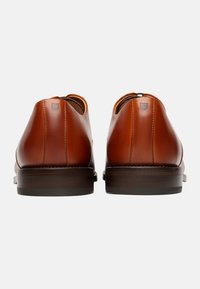 Chaussures en cuir marron avec une finition lisse, des talons arrondis et des coutures ton sur ton. Les semelles sont sombres et légèrement texturées pour une meilleure adhérence.