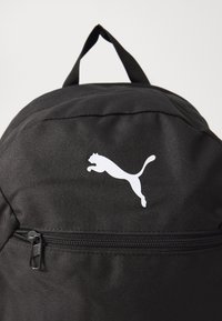 Zaino nero realizzato in tessuto resistente, con una tasca frontale con zip e un logo Puma bianco. Include un anello superiore per il trasporto.