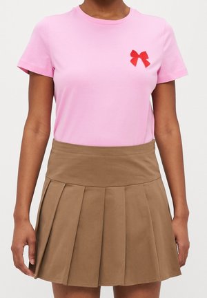 Vrouw die een roze shirt met korte mouwen draagt, met een kleine rode strik op de borst en een hooggetailleerde bruine geplooide minirok.