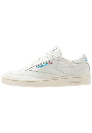 Baskets basses en cuir blanc Reebok avec logo bleu et détail du drapeau britannique sur le côté, semelle texturée et fermeture à lacets.