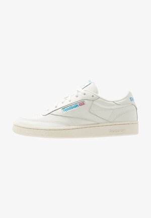 Baskets basses en cuir blanc Reebok avec logo bleu et détail du drapeau britannique sur le côté, semelle texturée et fermeture à lacets.