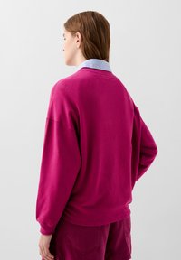 Fuchsia oversized sweatshirt met lange mouwen, geribbelde halslijn en een ontspannen pasvorm, gecombineerd met een lichtblauw shirt met kraag eronder.