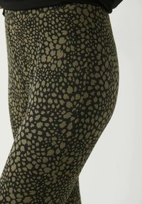 Leggings noirs avec un motif abstrait texturé en vert olive foncé. Ils présentent un tissu lisse et une coupe ajustée.