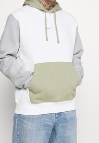 Hoodie med vit kropp, grå ärmar och en grön ficka. Tillverkad av mjukt material med dragsnören och en liten logotyp.