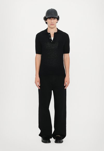 Schwarz gestricktes Poloshirt mit kurzen Ärmeln, zwei Knöpfe am Kragen. Kombiniert mit weiten schwarzen Hosen und schwarzen Schuhen. Schwarze Kappe auf dem Kopf.