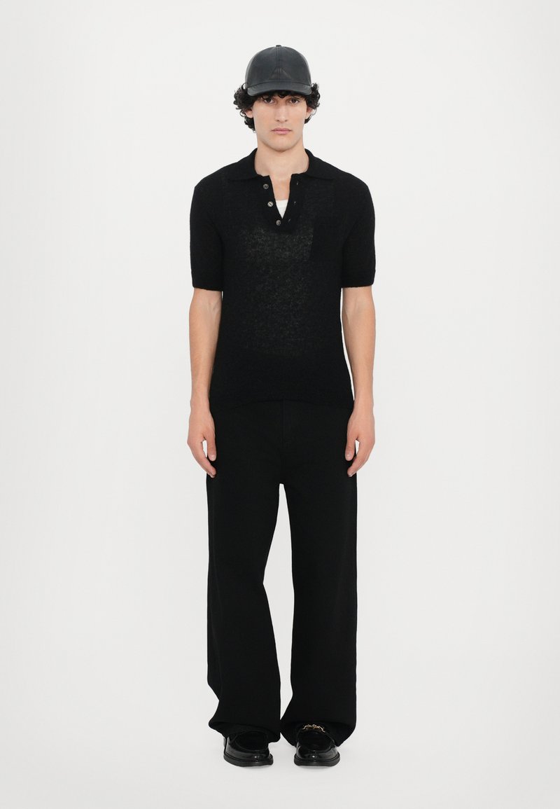 Schwarz gestricktes Poloshirt mit kurzen Ärmeln, zwei Knöpfe am Kragen. Kombiniert mit weiten schwarzen Hosen und schwarzen Schuhen. Schwarze Kappe auf dem Kopf.