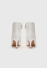 Paire de bottines blanches à talons hauts avec une texture florale subtile et des fermetures éclair à l'arrière, vues de dos sur un fond uni.