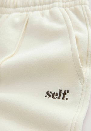 Sonnencremefarbene Jogginghose aus Stoff mit weicher Textur, elastischem Bund, Seitentaschen und "self." in Braun bestickt.
