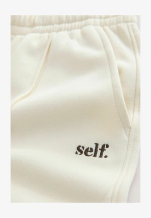 Svedbukser i cremefarvet stof med blød tekstur, elastisk taljebånd, sidelommer og "self." broderet i brun.