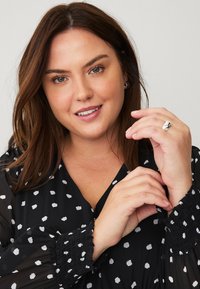 Zwarte blouse met wit stippenpatroon, doorschijnende stof, geplooide mouwen. Model toont sieraden op haar handen en glimlacht naar de camera.