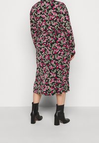 Frau in einem schwarzen Midikleid mit pinkem Blumenmuster und schwarzen, high-heeled Stiefeletten, die vor einem schlichten hellen Hintergrund steht.