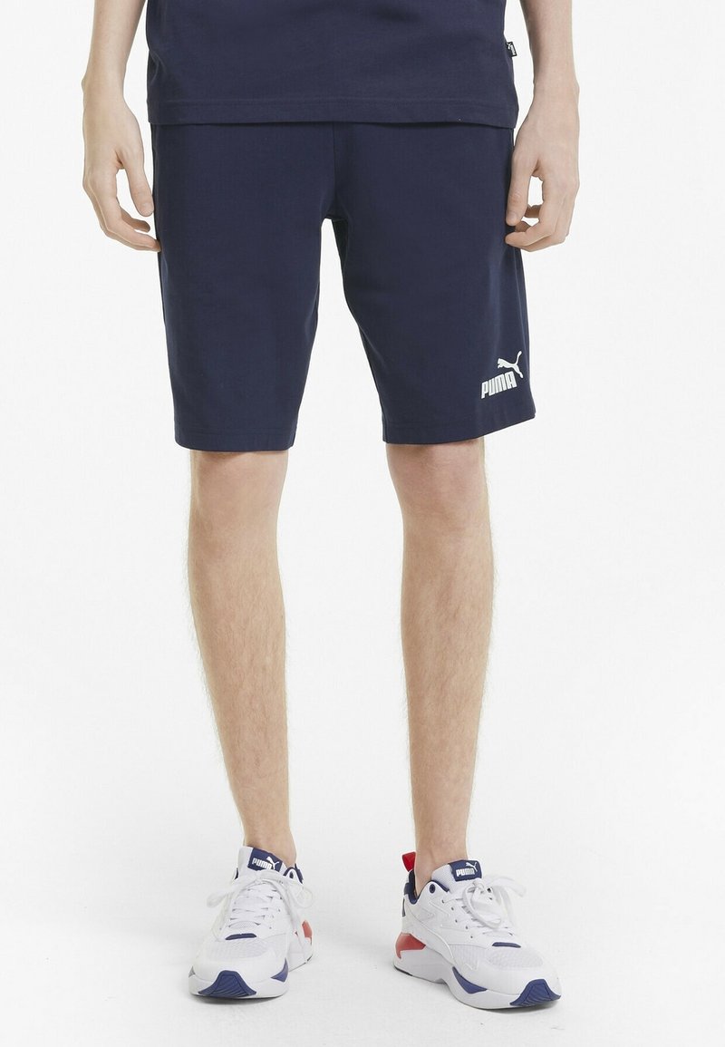 Jambes masculines portant un short Puma bleu marine et des baskets blanches Puma sur fond blanc.