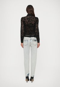 Woman wearing a sheer black floral lace long-sleeve top, light washed straight-leg jeans, and black high-heeled shoes, facing away.