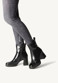 Stivaletti alla caviglia in pelle verniciata nera con tacco spesso, dettaglio con zip laterale e finitura liscia e lucida. Abbinati a jeans skinny grigio scuro.