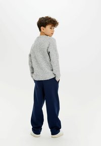 Maglione grigio chiaro a maglia, pantaloni blu navy larghi e sneakers bianche; bambino in piedi con le mani nelle tasche, visto da dietro.