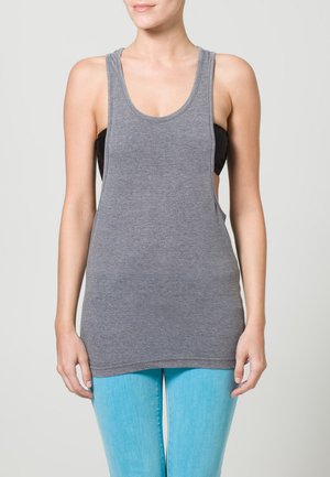 Jeune femme portant un débardeur gris ample sur un soutien-gorge de sport noir et un jean bleu clair, se tenant devant un fond neutre uniforme.