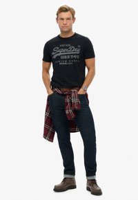 Camisa negra con texto de logo gris, vaqueros azul oscuro y botas de cuero marrón con cordones. Una camisa de cuadros roja está atada en la cintura.