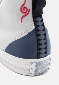 Κοντινό πλάνο της φτέρνας ενός αθλητικού παπουτσιού σε λευκό, ναυτικό μπλέ και μαύρο χρώμα, με κόκκινο κεντητό σχέδιο σπυροειδούς και μεταλλική πλάκα με εγχαραγμένο συμβολισμό σπείρας.