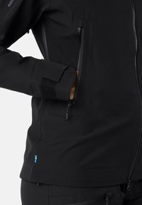 Giacca impermeabile nera con colletto alto, tasche laterali con zip e polsini texture con chiusura in Velcro. Dettaglio della piccola bandiera svedese.