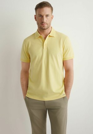 Mężczyzna stoi z rękami w kieszeniach, ubrany w żółtą koszulkę polo z krótkim rękawem oraz beżowe spodnie, na jednolitym tle.