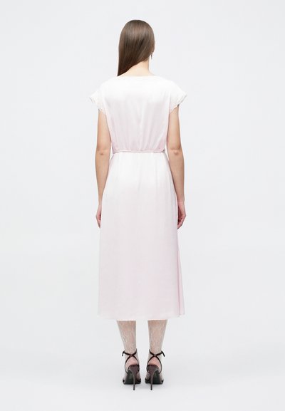 Γυναίκα που φοράει ροζ ανοιχτό midi φόρεμα με δαντελωτά μανίκια τύπου καπέλο, κολάν με σχέδια και μαύρες ψηλοτάκουνες σανδάλες, φαινομένη από πίσω.