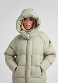 Donna che indossa un cappotto invernale imbottito di colore beige chiaro con un ampio cappuccio, con le mani nelle tasche, rivolta in avanti su uno sfondo neutro.