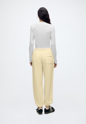 Femme aux longs cheveux foncés portant un top blanc à manches longues, un pantalon de survêtement beige avec une poche arrière et des baskets noires, debout de dos sur fond blanc.