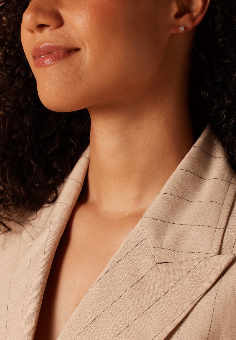 Detailaufnahme einer Person, die ein beiges, gestreiftes Blazer trägt. Der Stoff hat eine strukturierte Oberfläche, und kleine Steckerohrringe sind im Ohr sichtbar.