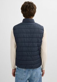 Chaleco acolchado en color navy con cuello alto, relleno acolchado y costuras horizontales, combinado con una camiseta de manga larga de color claro y jeans.