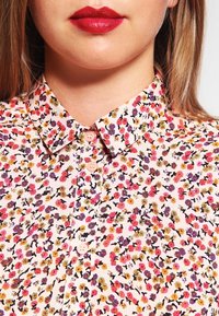 Donna che indossa una camicia a fiori con bottoni e colletto a punta, truccata con rossetto rosso brillante, vista dal mento fino alla parte superiore del petto.