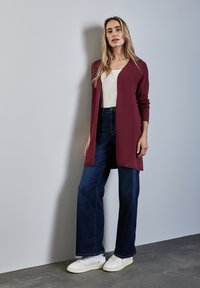 Bordeauxfarbener langer Cardigan über einem weißen Oberteil, kombiniert mit weit geschnittenen blauen Jeans und weißen Sneakers. Glatter Stoff und lässige Passform.
