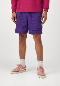 Lilla cargo-shorts med to sidelommer, en afslappet pasform og en glat tekstur, parret med lyserøde sneakers og hvide ankelstrømper.