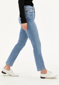 Persoon die loopt in lichtblauwe slim-fit jeans, een zwart lang-sleeve top en witte veterschoenen op een witte achtergrond.