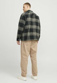 Cămașă din flanel în carouri negre și crem, cu guler în vârf, mansete cu nasturi și yoke pe spate; purtată cu pantaloni bej cu croială lejeră și adidași albi.